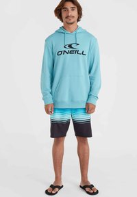 Felpa blu chiaro con logo O'Neill nero, abbinata a pantaloni da bagno a righe blu e neri. Indossata con ciabatte nere.