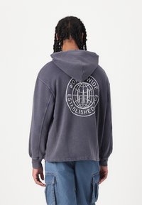 Hoodie din material moale, texturat, de culoare gri închis, având un logo mare alb pe spate, glugă și manșete ribbate.
