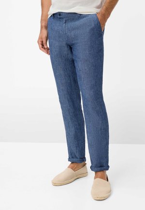 Mand iført tætsiddende blå linnedbukser med op rullede manchetter, beige slip-on sko og hvid top, stående med hænderne i lommerne.