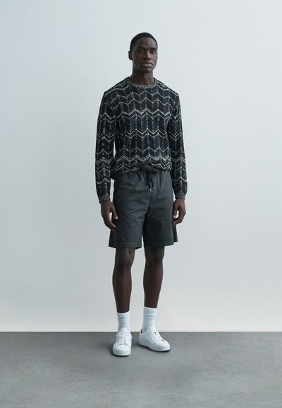 Sweter w czarno-białe geometryczne wzory na drutach, szare krótkie spodenki, białe trampki z prążkowanymi skarpetkami, stojące na jasnoszarym tle.