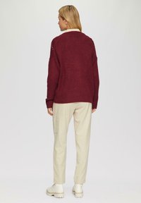 Burgunderfarbener Strickpullover mit lockerer Passform, geripptem Saum und überschnittenen Schultern; kombiniert mit cremefarbenen, maßgeschneiderten Hosen und hellen Schuhen.