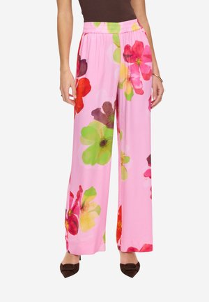 Donna che indossa pantaloni rosa a gamba larga a vita alta con grandi stampe floreali colorate, abbinati a tacchi marroni con punta appuntita.