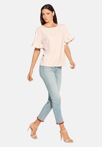 Helles rotes Oberteil mit Rüschensäumen, kombiniert mit hellblauen knöchellangen Jeans, die rote Akzente an den Säumen haben, und beigen, schnürbaren High Heels.