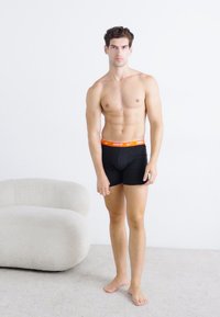 Black boxer briefs with an orange Nike logo waistband, fitted design, smooth texture, worn by a standing male model against a light background.