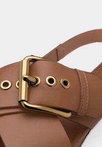 Λουρί σανδαλιού από ταμπά δέρμα με αγκράφα και μεταλλικά οφθαλμός σε χρυσό, κοντινή λήψη που δείχνει την υφή και τις ραφές.