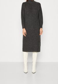 Donna in un lungo abito in maglia grigia scura con collo alto, abbinato a stivali bianchi fino al ginocchio, che si trova davanti a uno sfondo chiaro e neutro.