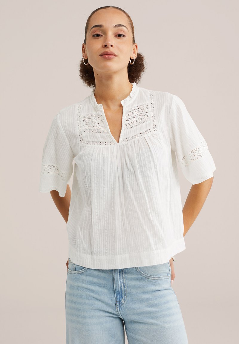 Femme aux cheveux bouclés portant des boucles d'oreilles créoles, un chemisier blanc texturé avec des détails en dentelle et un jean bleu clair, debout devant un fond uni.