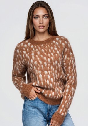 Une femme pose devant un fond uni. Elle porte un pull marron à pois crème et un jean bleu. Elle a une main dans la poche.