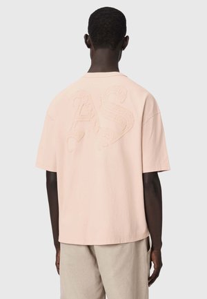 Mężczyzna widziany od tyłu, ubrany w luźną bladoróżową koszulkę z wypukłymi fakturowanymi literami „AS” na plecach oraz beżowe spodnie na jednolitym tle.