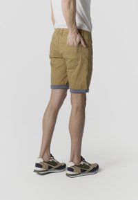 Férfi áll, felhajtott khaki sorttal, fehér pólóban, bézs, fekete és zöld sportcipőben, kezét a hátsó zsebében tartva.