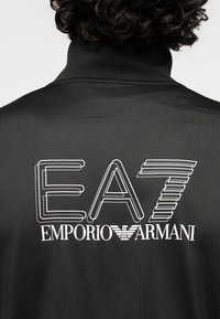 Μαύρο αθλητικό μπουφάν με ψηλό γιακά, υφασμένο ύφασμα και μεγάλο λευκό λογότυπο με το "EA7" πάνω από το "EMPORIO ARMANI" στην πλάτη.