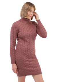 Žena s krátkými blonďatými vlasy, oblečená v mauve svetr šaty s pleteným vzorem a rolákem, se dotýká svých vlasů a jemně se usmívá.