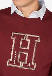 Bordowy sweatshirt z dużym haftowanym "H" w beżowym ściegu, z napisem "HARPER" poniżej. Miękki materiał bawełniany, okrągły dekolt.