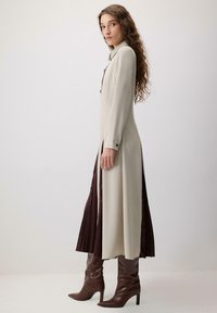 Lang, beige kjole med sideslids, knapdetaljer og krave; parret med brune, knæhøje, spidse støvler. Glat tekstur.