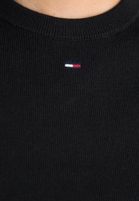 Svart strikket genser med ribbestruktur, med en liten brodert logo i blått, hvitt og rødt nær kragen.