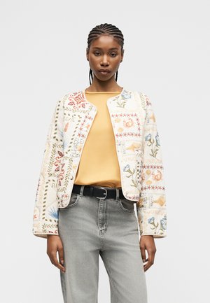 Kvinde med flettet hår iført en cremefarvet jakke med mønster over en gul top og lysegrå jeans med sort bælte, stående mod en ensfarvet baggrund.