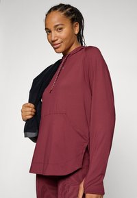 Mörkröd långärmad hoodie med en framficka, sidospår och en justerbar snörning på huvan; mjuk textur och avslappnad passform.