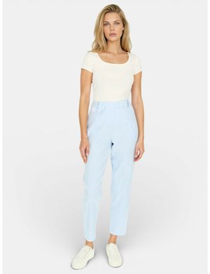 Kobieta stoi, ubrana w biały krótki rękaw, jasnoniebieskie spodnie z wysokim stanem oraz białe trampki na jednolitym jasnym tle.