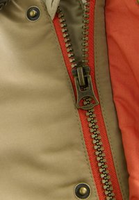 Dettaglio di una giacca khaki con zip, caratterizzata da un nastro zip rosso, una chiusura in ottone e una texture di tessuto liscia con una fodera arancione in contrasto.