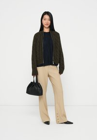 Olivgrüner, kabelgestrickter Bomberjacke, marinebluse mit Falten, beigen weiten Hosen mit Knöpfen und einer kleinen schwarzen Handtasche. Schwarze spitze Schuhe.