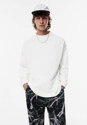 White long-sleeve shirt with a relaxed fit, paired with black patterned pants featuring teal and grey details. A black-and-white cap completes the outfit.