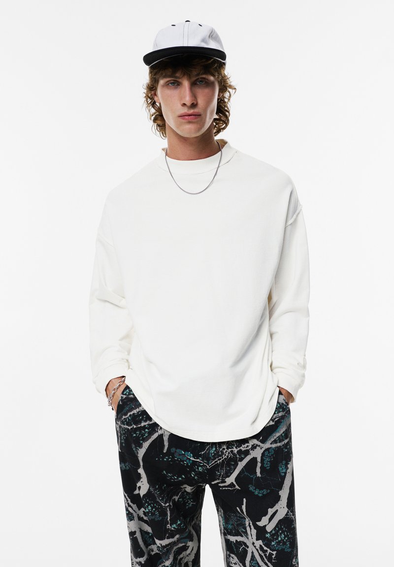 Chemise blanche à manches longues avec une coupe décontractée, assortie à un pantalon noir à motifs avec des détails turquoise et gris. Une casquette noir et blanc complète la tenue.