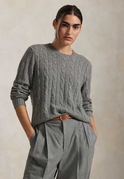 Frau mit dunklen Haaren, die einen grauen Zopfstrickpullover, graue plissierte Hosen und einen braunen Gürtel trägt, steht mit den Händen in den Taschen.
