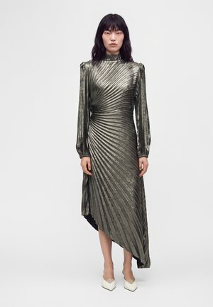 Femme portant une robe métallisée à manches longues avec des plis diagonaux et un ourlet asymétrique, associée à des talons blancs à bout pointu.