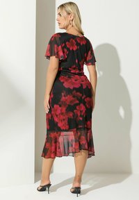 Robe midi noire à fleurs avec des fleurs rouges et roses, en tissu léger, avec des manches flutter et un ourlet évasé. Portée avec des mules à talons noirs.
