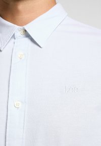 Camisa de color azul claro con cuello, hecha de un tejido texturizado, que presenta dos botones blancos y un logotipo bordado de "Lee" en el pecho.