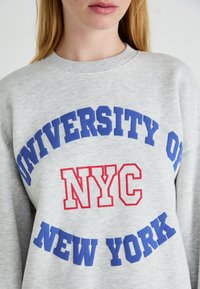 Close-up de uma pessoa a usar um moletom cinza claro com "Universidade de Nova Iorque" em azul e "NYC" em letras vermelhas na parte da frente.