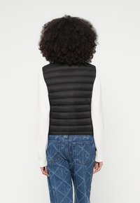 Μαύρο γιλέκο puffer με οριζόντια ραφή, ελαφρύ σχέδιο, φορεμένο πάνω από λευκό μπλουζάκι με μακριά μανίκια, συνδυασμένο με μπλε τζιν με σχέδια.