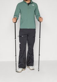 Zielona kurtka z zamkiem błyskawicznym i dwoma bocznymi kieszeniami, w połączeniu z czarnymi spodniami z zamkami. Trzyma dwa kijki trekkingowe, nosi jasne buty do wędrówek.