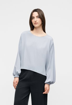 Kobieta o prostych brązowych włosach, ubrana w luźną, jasnoszarą bluzkę z długim rękawem z elastycznymi mankietami oraz czarne spodnie z wysokim stanem, stoi na tle białego tła.