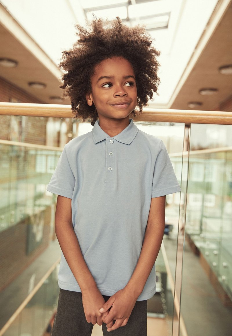 Göndör hajú gyermek világoskék galléros pólót viselve, belül áll az üvegkorlát mellett, enyhén jobbra néz, lágy mosollyal az arcán.