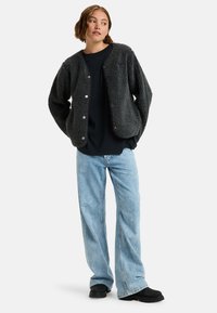 Grijze, fluffy, gestructureerde jas met drukknoopsluiting, gedragen over een zwart shirt. Gecombineerd met lichtblauwe, loszittende jeans en zwarte schoenen.