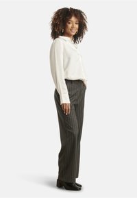White button-up shirt, gray pinstripe trousers, black block-heeled shoes; model stands sideways with curly hair and hands in pockets.