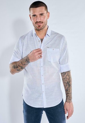 Mann mit kurzen Haaren und Tätowierungen an den Armen, trägt ein hellblau kariertes Hemd und dunkle Jeans, richtet den Hemdkragen, steht.