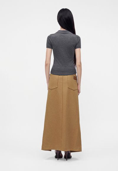 Femme aux longs cheveux foncés portant un haut gris ajusté à manches courtes et une jupe longue camel taille haute avec poches, debout, de dos.