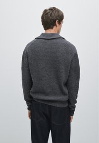 Pull gris en tricot avec texture côtelée, col large et manches longues. Porté par-dessus un jean foncé, vu de dos sur un fond uni.