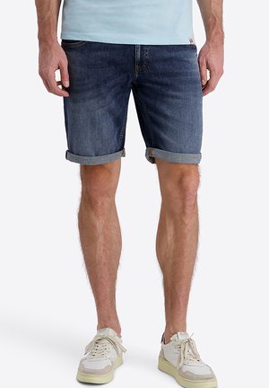 Man wearing rolled-up dark denim shorts and white sneakers, standing with hands relaxed by his sides.