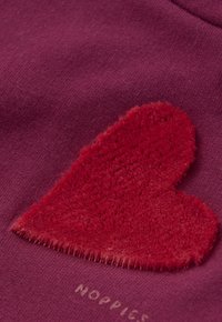 Rood harig hart appliqué genaaid op een paarse stoffen achtergrond met gedeeltelijk zichtbare lichte tekst die "NOPPICS" zegt.