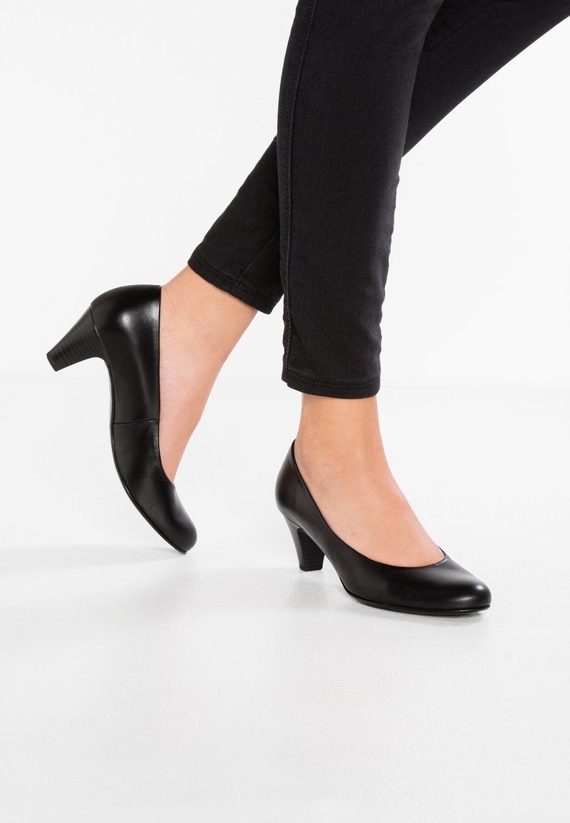 Personne portant des escarpins en cuir noirs à talons moyens, associés à un pantalon skinny noir, debout devant un fond blanc.