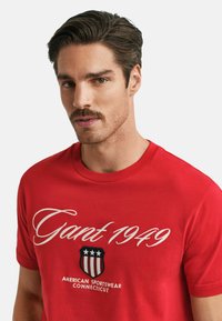 Rotes Baumwoll-T-Shirt mit weißer Schrift "Gant 1949" und einem Wappen-Logo mit Sternen und Streifen. Rundhalsausschnitt und kurze Ärmel.