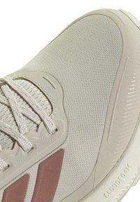 Zapatilla deportiva beige con parte superior de malla texturizada, cordones claros y un acento burdeos. Presenta una suela pronunciada etiquetada como "CLOUDFOAM."