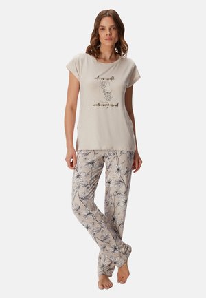 Femme portant un haut beige à manches courtes avec imprimé floral et texte, associé à un pantalon de pyjama beige à motif floral, debout pieds nus.