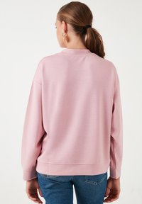 Felpa rosa con scollo rotondo e maniche lunghe, realizzata in tessuto liscio. L'orlo è leggermente accorciato, abbinata a jeans blu aderenti.