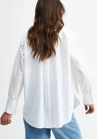 Chemise blanche à manches longues avec une coupe décontractée, dotée d'un ourlet arrondi, de manches tombantes et de poignets boutonnés, associée à un jean bleu texturé.