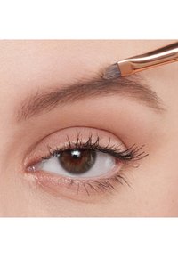 Zbliżenie brązowego oka z długimi rzęsami i delikatnie przyciemnionymi powiekami, pokazujące rękę nakładającą produkt do brwi różowozłotym pędzelkiem.