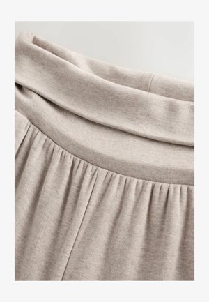 Weicher beiger Stoff mit einem gerafften Nahtdetail und einem breiten, gefalteten Bund, der die Textur und die Nähte der Kleidung hervorhebt.